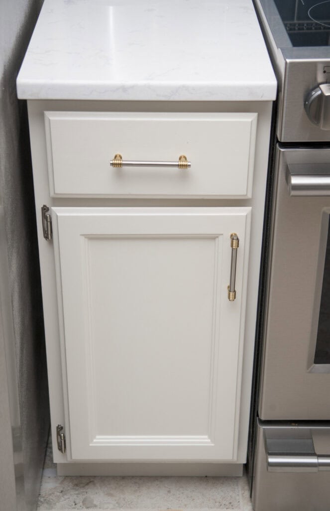 A narrow white kitchen cabinet with gold handles, featuring one drawer on top and a door below, situated between a wall and a stainless steel oven.