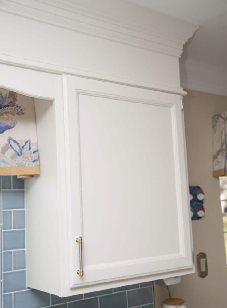 A white kitchen cabinet with a gold handle, mounted above a blue tiled backsplash. Floral-patterned window valances are partially visible on both sides.