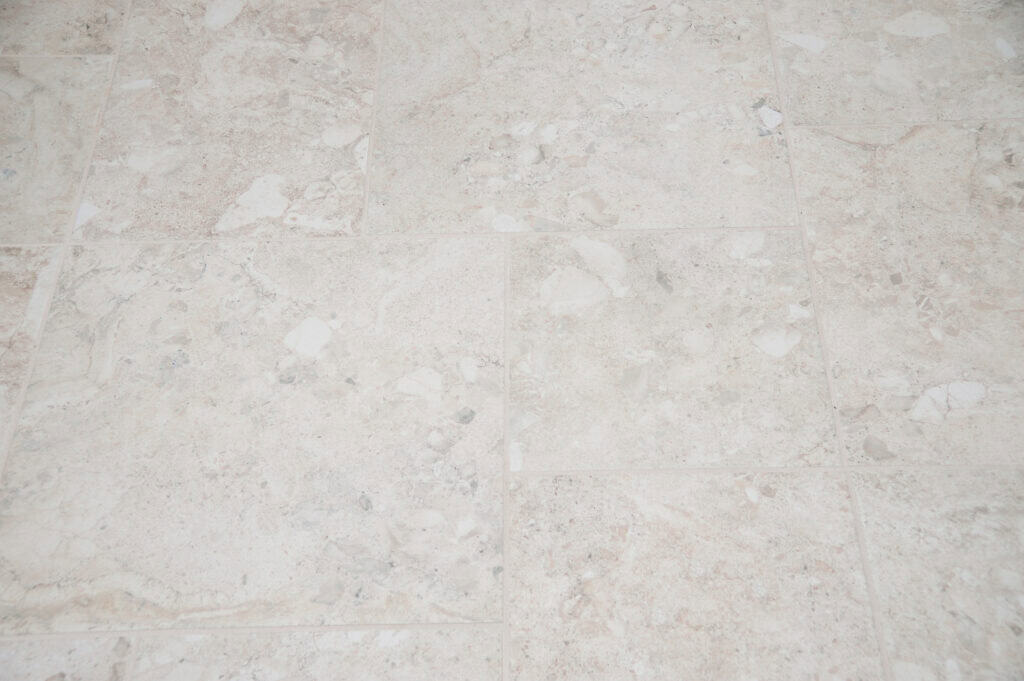 Close-up of light beige marble tiles with subtle natural patterns and specks of gray and cream, arranged in a grid pattern. The surface appears smooth and slightly reflective.