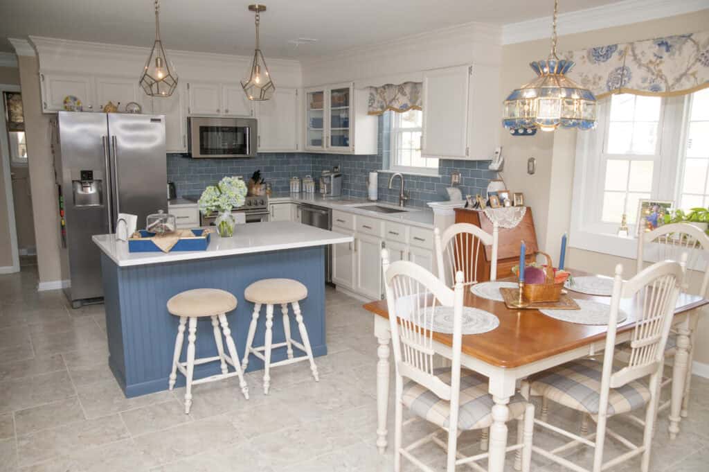 Bright kitchen with white cabinets, a blue island with two stools, stainless steel appliances, blue tile backsplash, and a dining table with four chairs, candles, and a fruit basket under large windows.