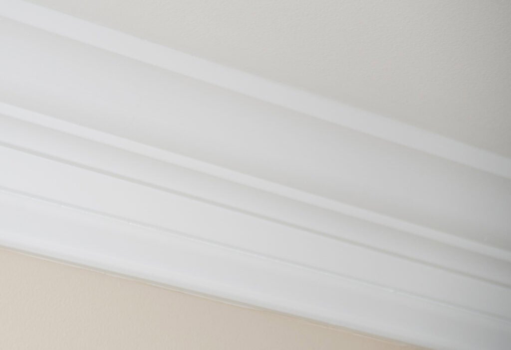 A close-up view of white crown molding where a light-colored wall meets a white ceiling, showcasing the architectural detail and smooth finish.