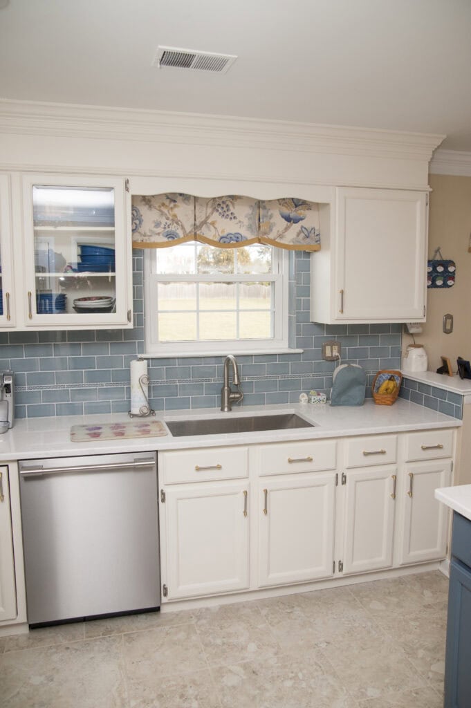 A clean kitchen with white cabinets, light blue tile backsplash, stainless steel dishwasher, single sink under a window with a floral valance, and neutral tile floor. Countertops hold paper towels, a toaster, and other small items.