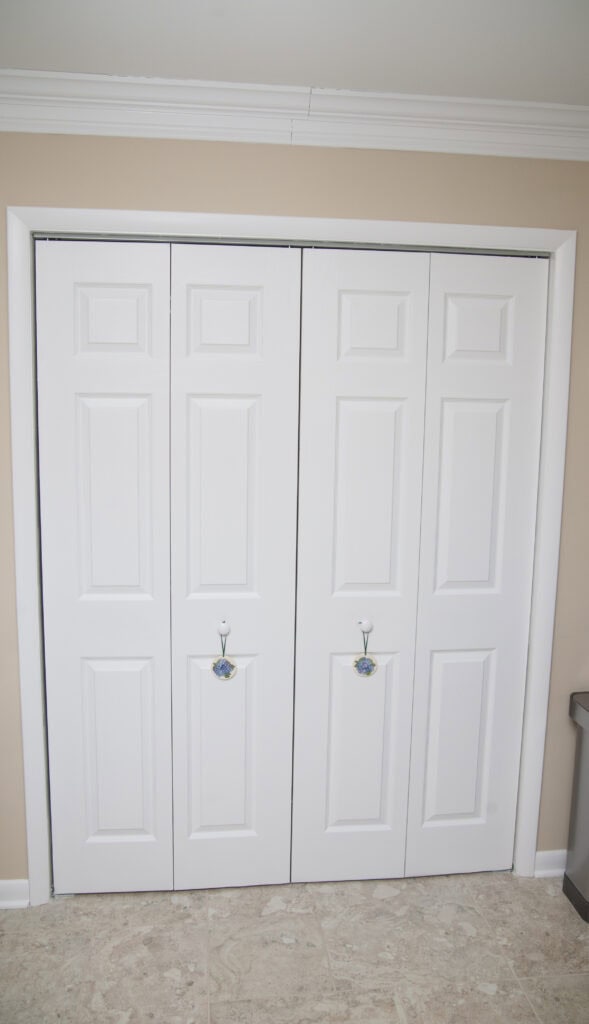 White double bi-fold closet doors with four raised panels each, set in a beige wall. Each door has a decorative hook with a colorful knob. The floor is light-colored tile, and the room has white crown molding.