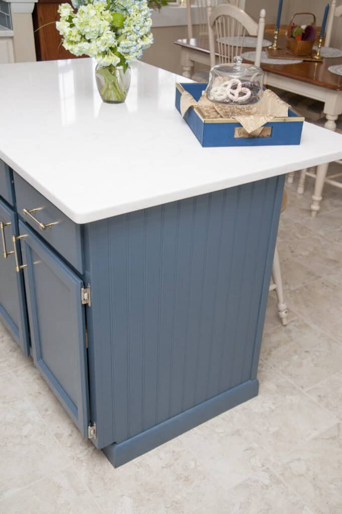A kitchen island with blue cabinets and a white countertop holds a clear vase with blue and green flowers and a tray with pastries on a beige tiled floor. Light-colored chairs and a table are in the background.