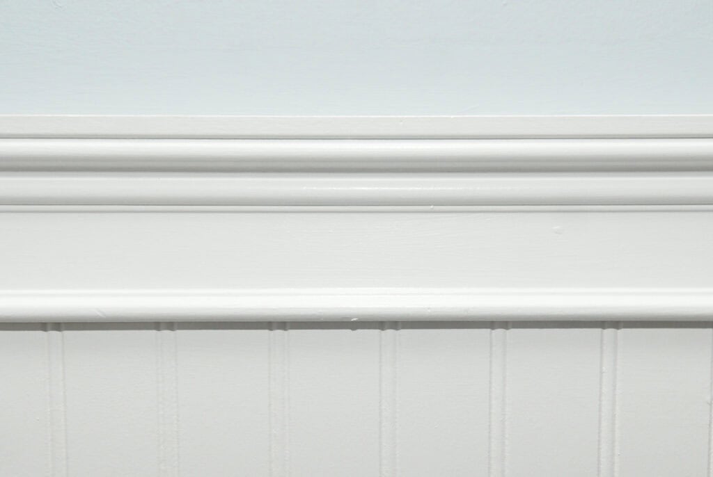 White-painted wainscoting with decorative trim against a light-colored wall; the paneling features vertical grooves below a smooth horizontal ledge and moulding.