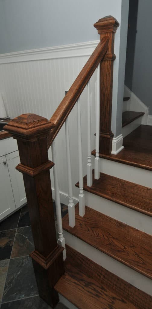 A wooden staircase with dark stained steps, matching newel posts and handrail, and white balusters. The stairway is adjacent to a white paneled wall and cabinet, with slate tile flooring visible.