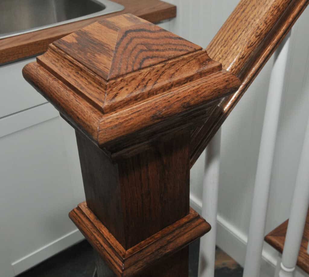 Close-up of a dark wood staircase newel post with decorative trim and grain patterns, next to white stair spindles and a kitchen counter with a sink in the background.