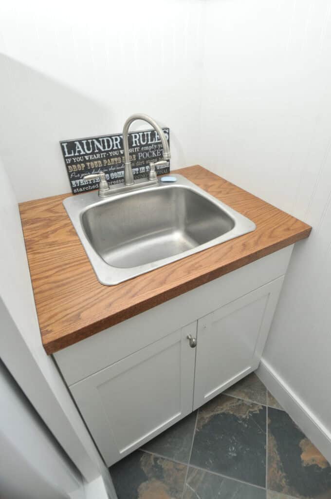 A small laundry room sink with a stainless steel basin, wooden countertop, white cabinet below, and a faucet. A decorative “Laundry Rules” sign is displayed at the back against white walls.