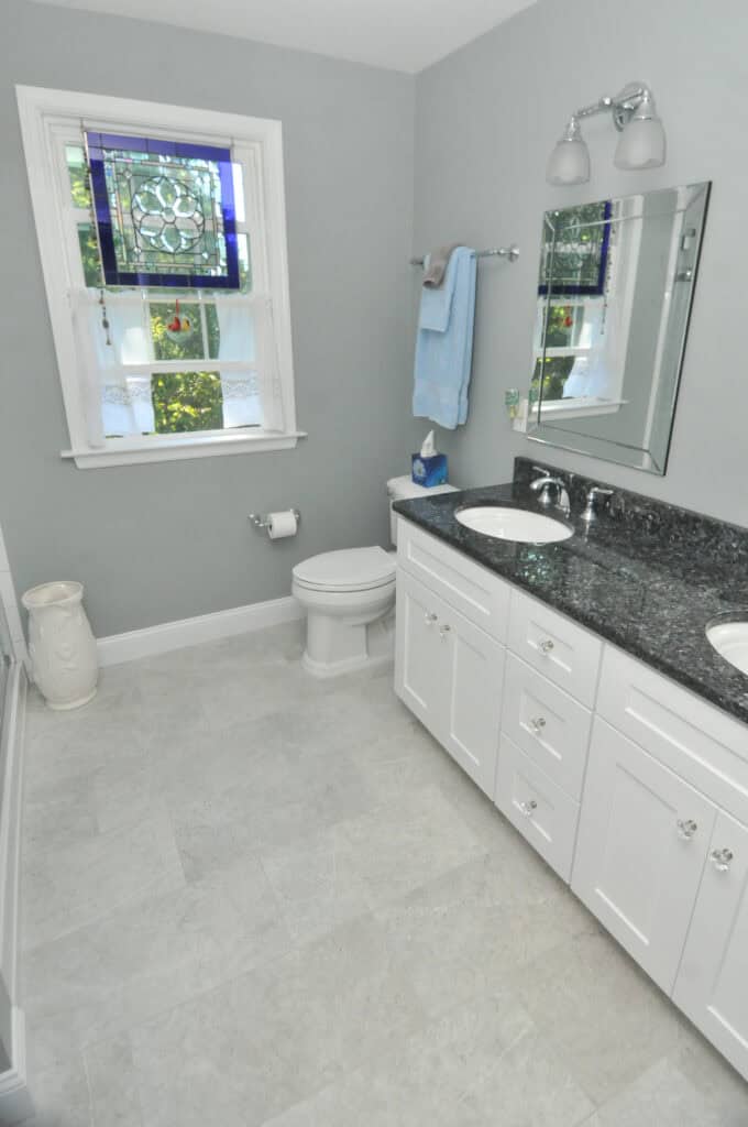 A bright, clean bathroom with a double-sink vanity, granite countertop, large mirror, toilet, and a window with a stained glass panel and sheer curtains. White cabinets and light gray walls and floor tiles.