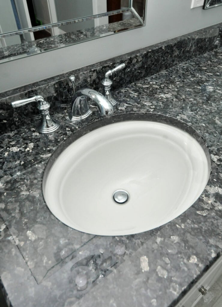 A white oval sink with a silver faucet and handles set in a dark marble countertop, seen from above. A wall mirror and a portion of the backsplash are visible in the background.
