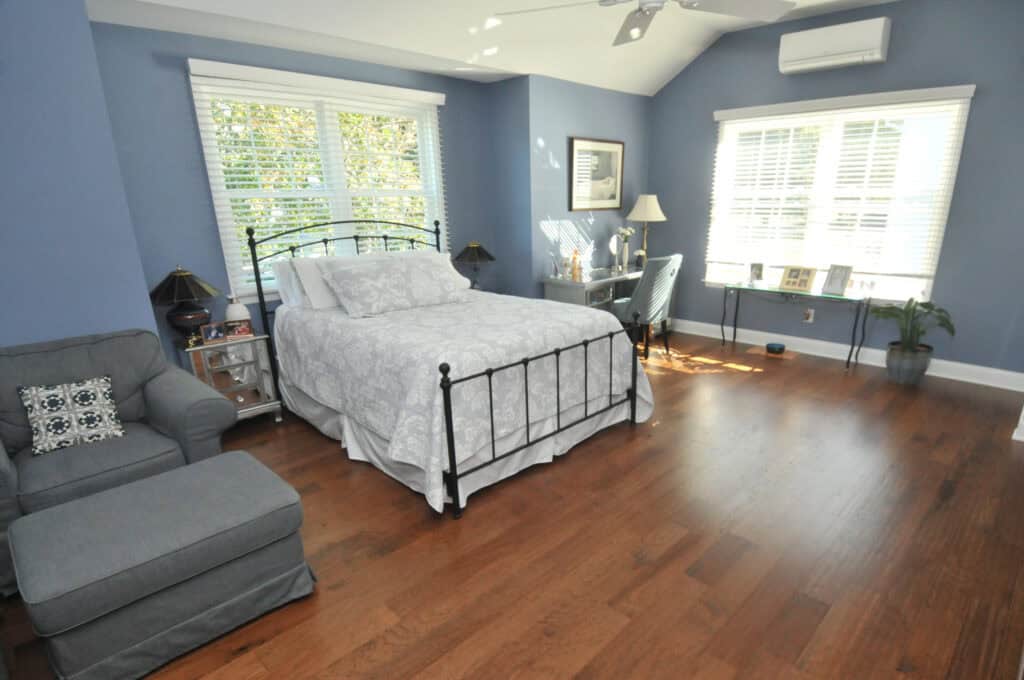 A spacious bedroom with blue walls, wood flooring, a metal-framed bed with white bedding, a gray armchair, a desk by the window, and sunlight streaming in through large windows with white blinds.