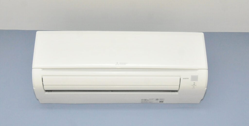 A white wall-mounted air conditioner unit attached to a light blue wall, seen from the front. The unit is turned off, and indicator lights are not illuminated.