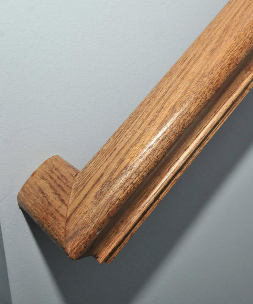 A close-up of a wooden handrail attached to a light gray wall, showing the end cap and grain detail of the polished wood.