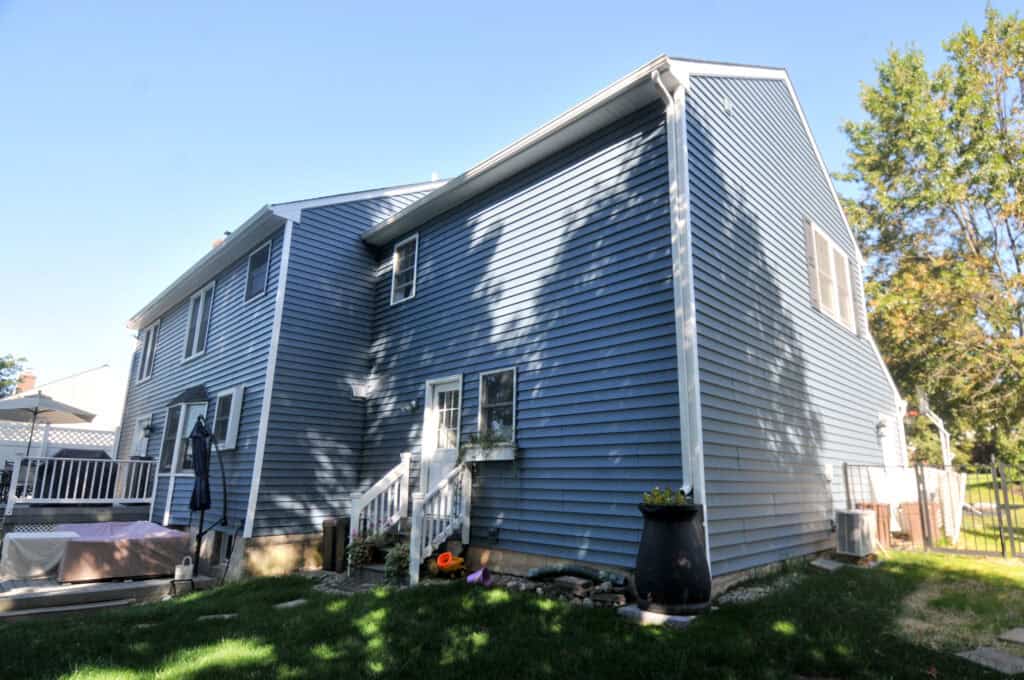 A two-story house with blue siding, white trim, and a small back porch. There are plants near the steps, a large black planter, and trees casting shadows on the house under a clear blue sky.