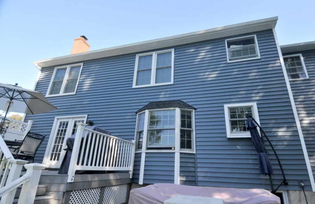 A two-story house with blue siding, white trim, a bay window, and a small deck with outdoor furniture and a staircase leading down.