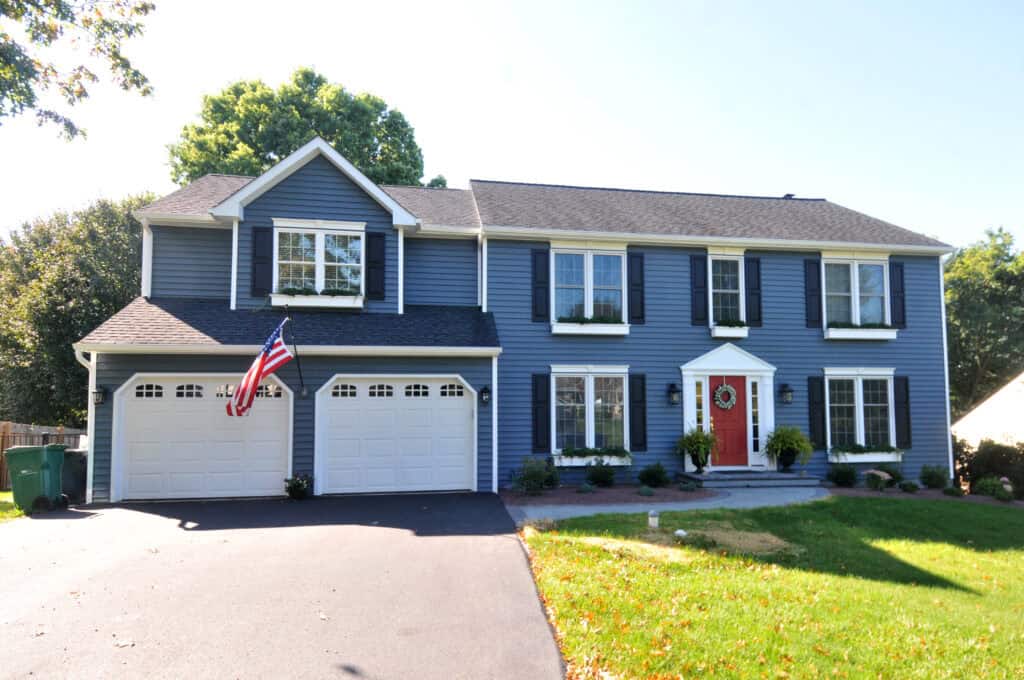 A two-story blue house with white trim, a red front door, two garage doors, an American flag, and a neatly landscaped front yard with green grass and potted plants.