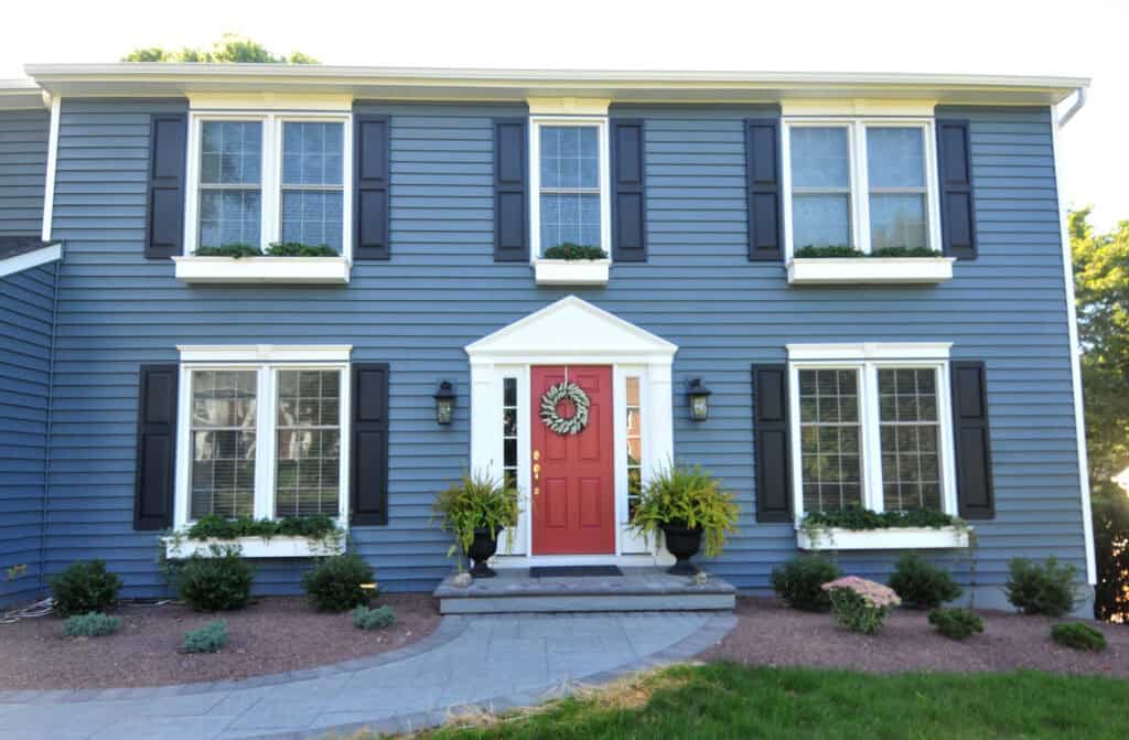 A two-story blue house with white trim, black shutters, and a red front door. The entrance is decorated with a wreath, two potted plants, and there are flower boxes under the windows. Small shrubs line the walkway.