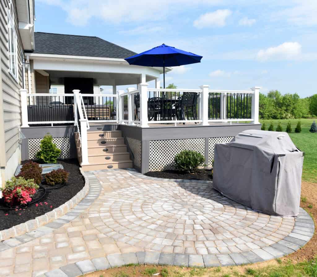 Backyard patio with a covered grill, circular stone pavers, landscaped garden beds, steps leading up to a raised deck with white railings, outdoor dining set, and a blue patio umbrella under a partly cloudy sky.
