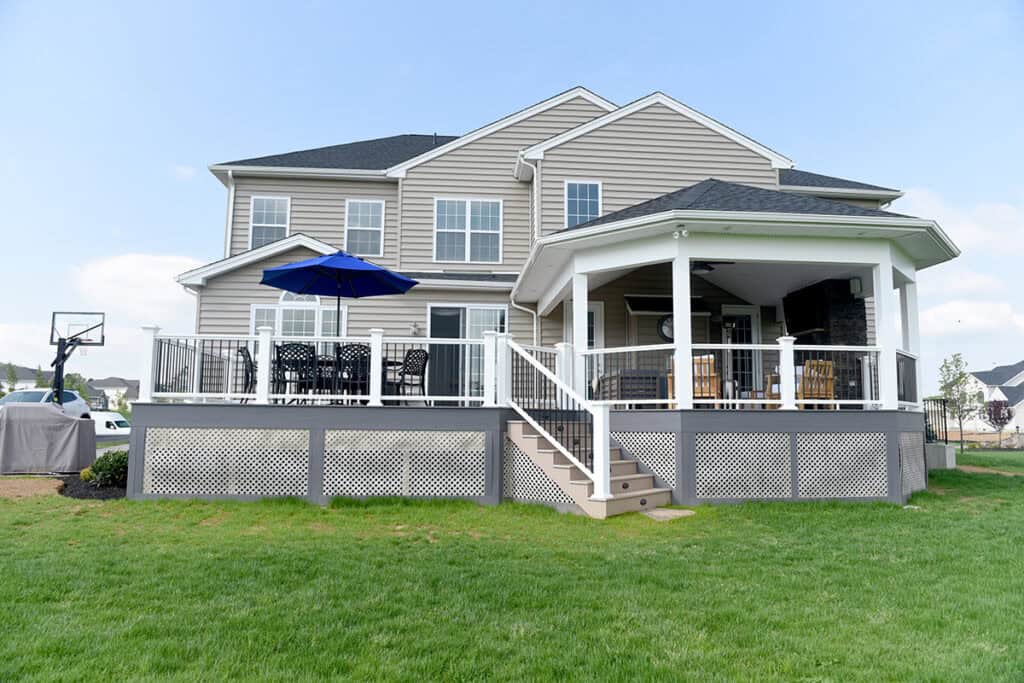 A two-story beige house with white trim has a large raised deck with a patio table, blue umbrella, and covered porch area, overlooking a green lawn under a partly cloudy sky.