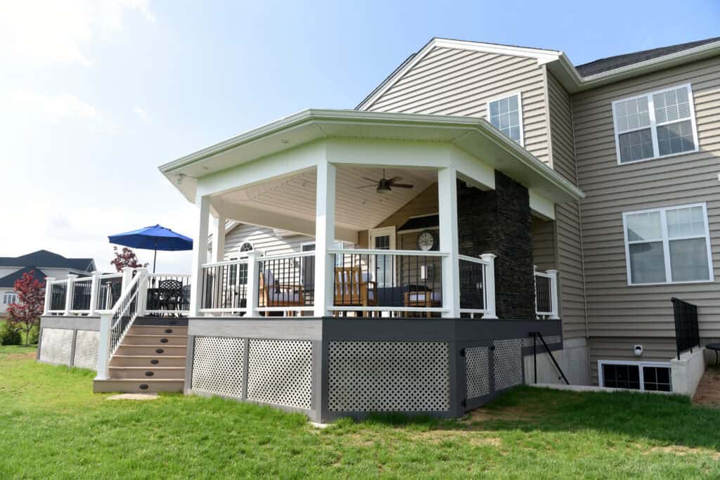A two-story beige house with a large covered porch featuring white railings, outdoor furniture, a stone accent wall, ceiling fan, and stairs leading down to a grassy yard. A blue umbrella shades part of the deck.