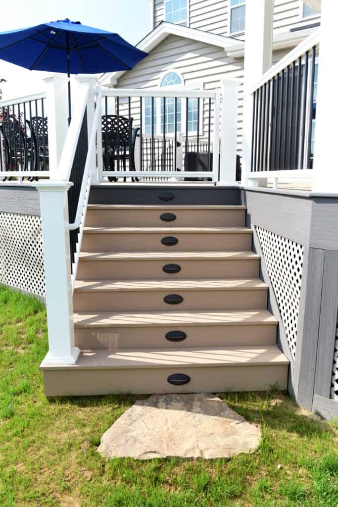 Outdoor steps lead up to a deck with white railings and black balusters. There are black oval accents on each riser. A patio table and blue umbrella are visible on the deck. Grass and a flat stone are at the base of the stairs.