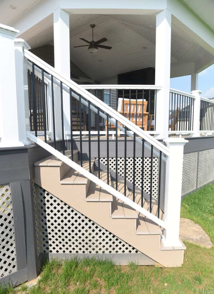 Outdoor staircase with beige steps and white railings leads up to a covered porch. The porch has white columns, a ceiling fan, wooden chairs, and decorative lattice skirting below. Grass and stone are at the base.