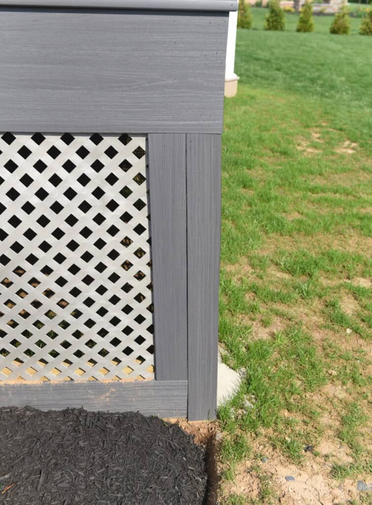A close-up of a gray lattice panel framed with matching gray wood, next to a patch of green grass and some mulch on the ground.