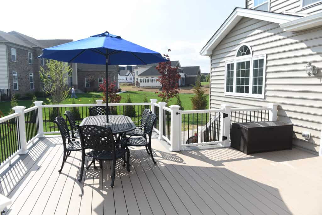 A spacious backyard deck with a black metal table and chairs under a blue patio umbrella, surrounded by white railing and overlooking green lawns and neighboring houses.