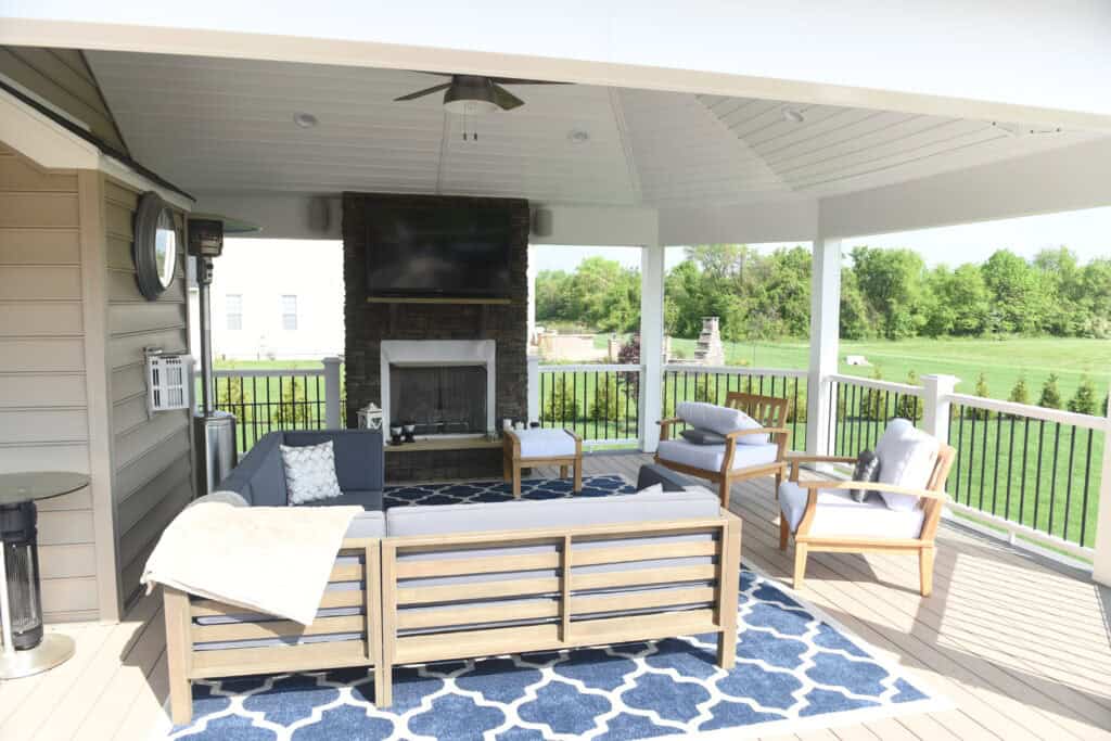 A covered outdoor patio features a gray sectional sofa, two armchairs, a fireplace with a TV above it, and a blue patterned rug. The area overlooks a green lawn with trees in the background.