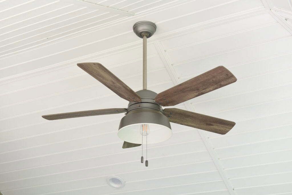 A ceiling fan with four wooden blades and a central light fixture hangs from a white, paneled ceiling. Two pull chains hang down from the fan.