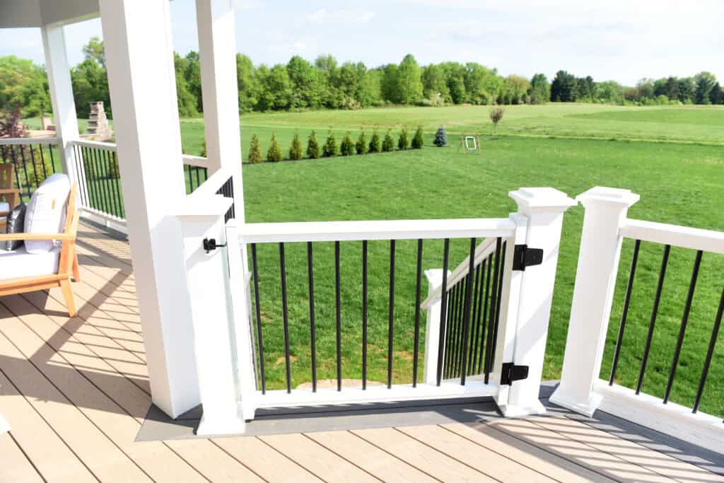 A white and black railing with a gate on a wooden deck overlooks a large, green lawn with trees and shrubs in the background under a clear sky.