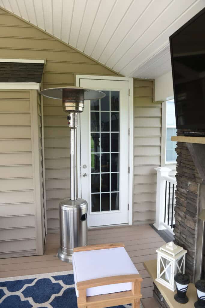 A patio area with a tall stainless steel outdoor heater, a glass-paneled door, beige siding, a TV, a small white lantern, and a chair with a blue and white rug on a wooden floor.