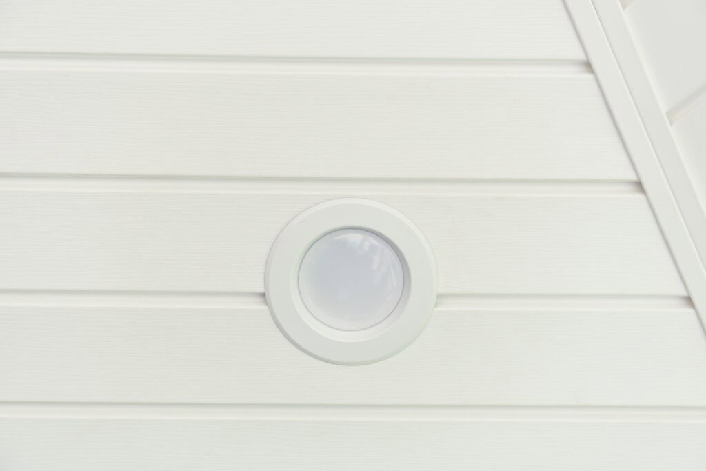 A round, white recessed ceiling light is mounted on white, horizontally paneled ceiling boards with a diagonal edge visible in the upper right corner.