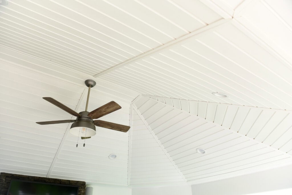 White paneled ceiling with a modern dark wood ceiling fan and several recessed lights. A portion of a TV and stone wall is visible at the bottom left corner of the image.