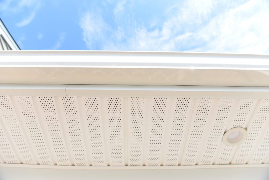 Close-up view of a white soffit with perforated panels and a recessed light, under the edge of a roof, with blue sky and clouds visible in the background.