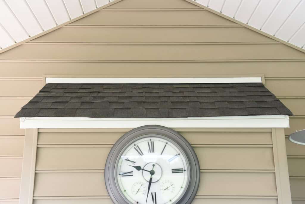 A round outdoor clock with Roman numerals is mounted on beige vinyl siding beneath a small gabled roof with gray shingles. The time on the clock is 4:42.