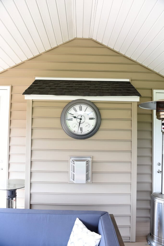 A large round wall clock with Roman numerals hangs on tan siding beneath a small roof structure. Below the clock is a silver vent. Part of a blue couch and a pillow are visible in the foreground.