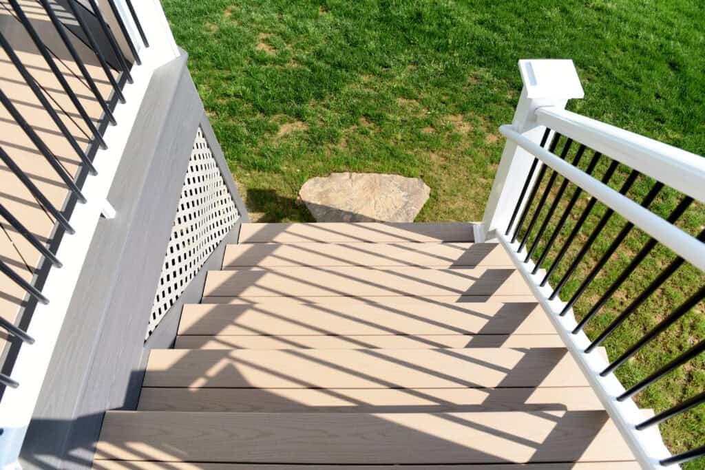 View looking down a set of beige outdoor stairs with white and black railings, casting shadows, leading to a grassy yard with a large stone at the bottom.