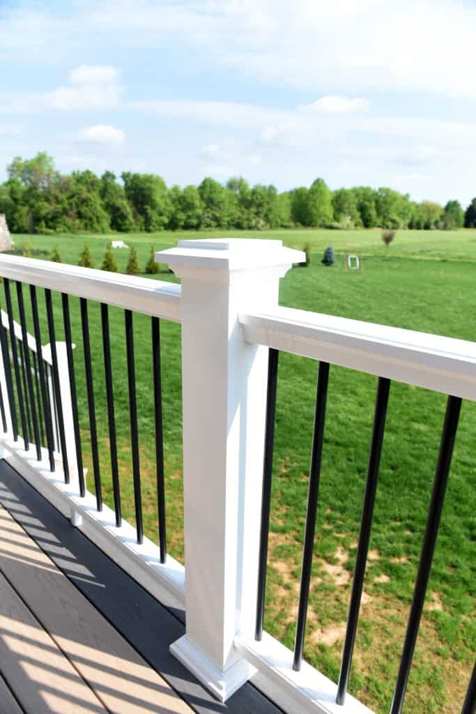 A close-up view of a white deck railing with black balusters, overlooking a green grassy lawn and trees under a partly cloudy sky.