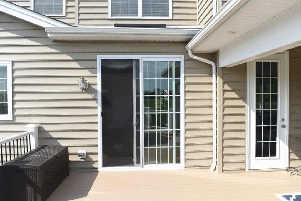 A beige house exterior with a sliding glass door featuring a screen, next to a white-framed glass door. The deck is light brown, and there is a black outdoor bench to the left.