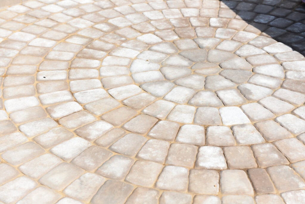 A circular pattern of light and dark stone pavers arranged in concentric rings on a paved surface, with a section in the upper right corner appearing darker, possibly shaded.