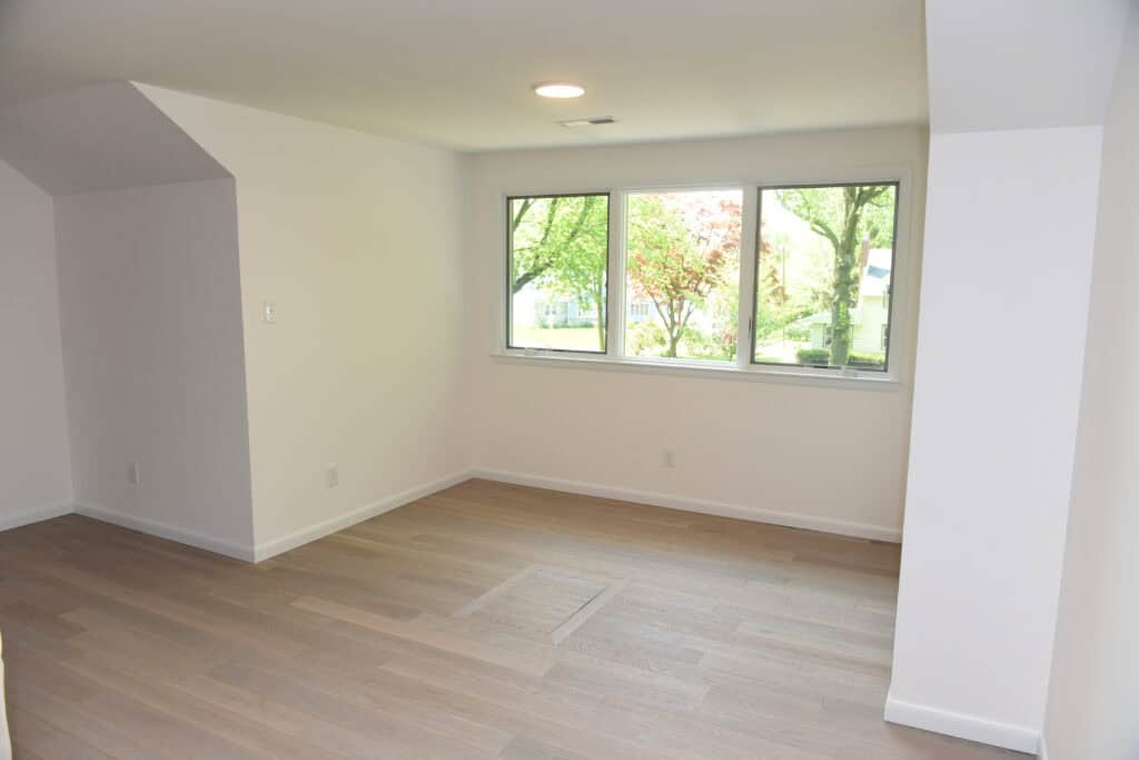 A bright, empty room with light wood floors, white walls, a recessed ceiling light, and a large window overlooking green trees and houses outside.