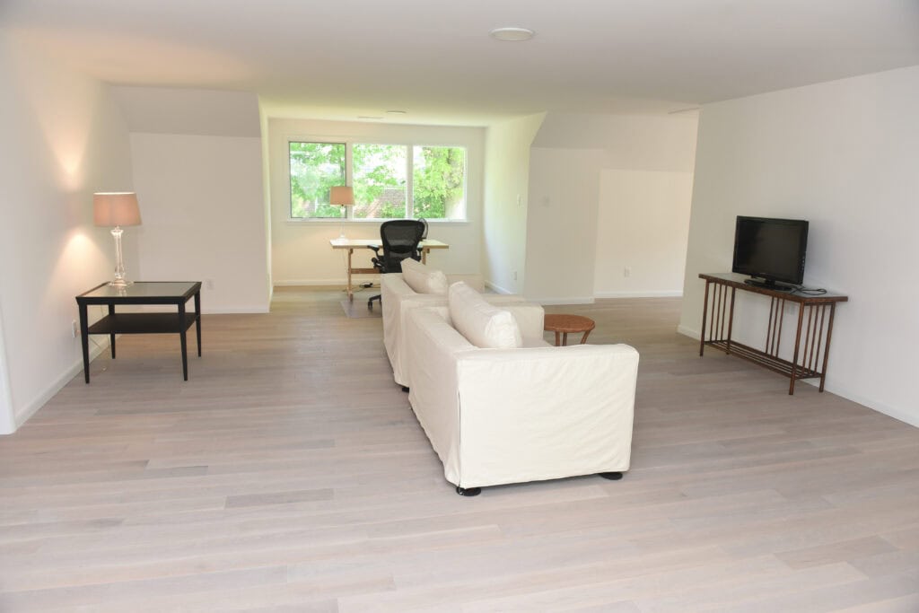A spacious, minimalist living room with light wood floors, white walls, a white sofa, TV on a stand, a small black table with a lamp, and a desk with an office chair by large windows showing greenery outside.