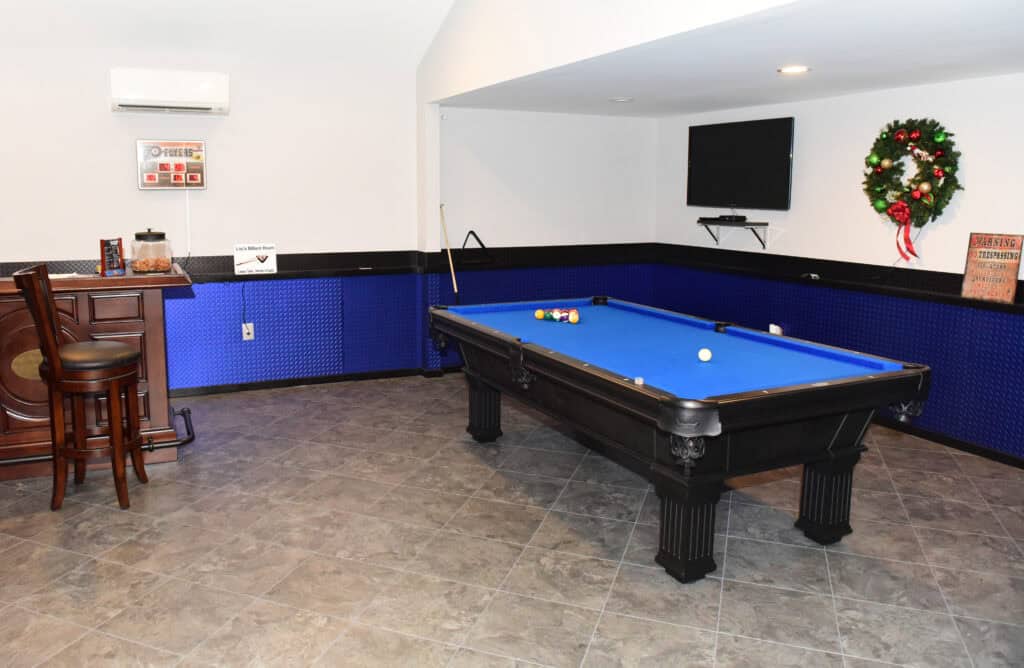 A room with a blue pool table, pool cues on the wall, a mounted TV, a holiday wreath, a small bar area with two chairs, and tiled floors. The walls have blue paneling and there is an air conditioner above the bar.
