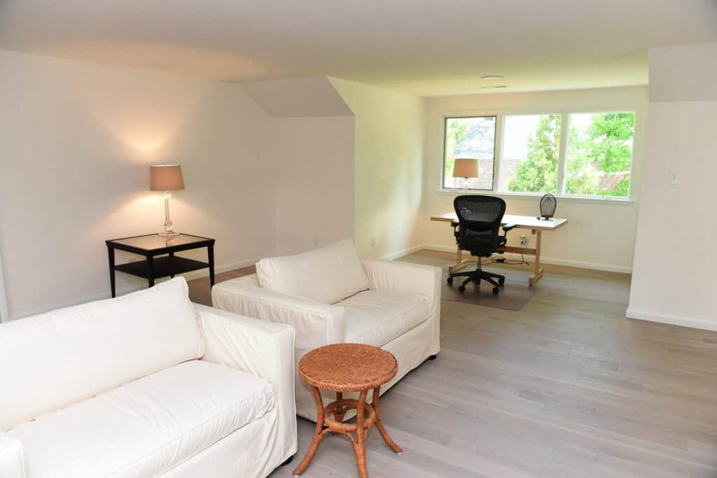 A bright, modern living space with light wood floors, white sofas, a wicker side table, a black desk with office chair by a window, and a table lamp on a black side table against the wall.