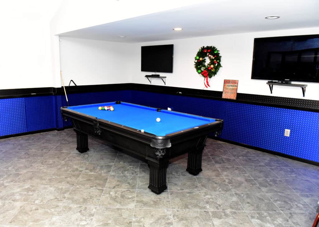 A blue pool table with several balls arranged on it is in a game room with blue and black walls, a cue stick, two wall-mounted TVs, a holiday wreath, and tiled flooring.