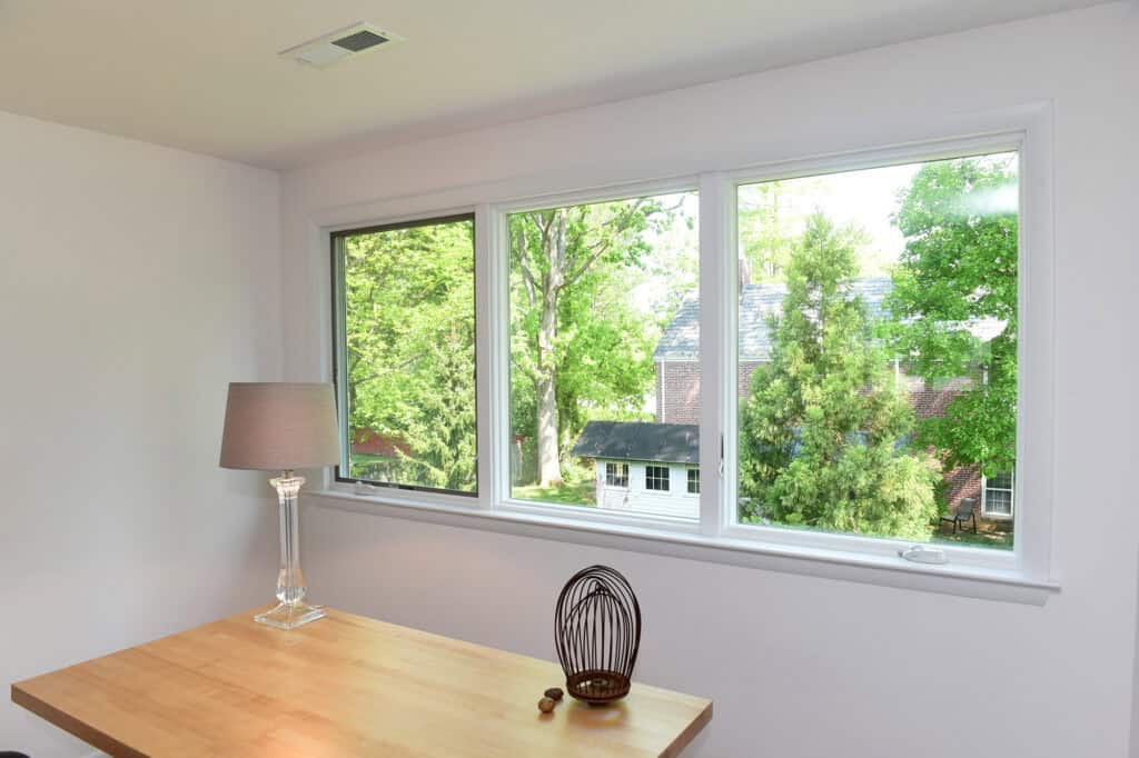 A bright room with a wooden desk, a clear lamp, and a decorative wire sculpture sits in front of large windows overlooking green trees and a house outside.