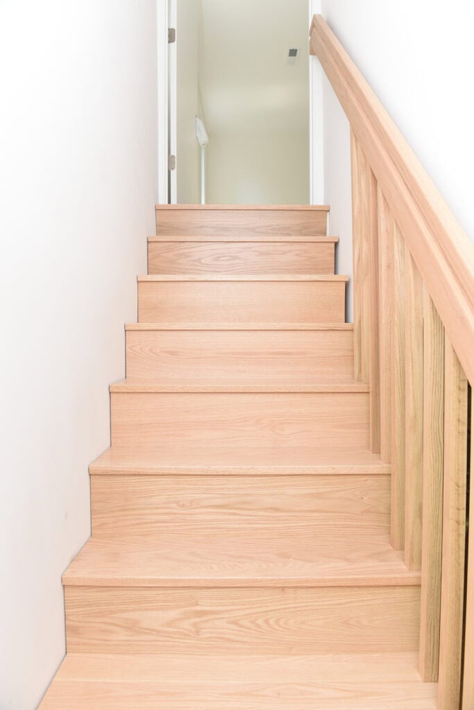 A set of light wood stairs with matching wooden railing on the right side, leading up to a white hallway with a closed door at the top. The area is brightly lit and the walls are plain white.