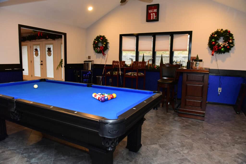 A game room with a blue pool table ready for play, bar stools, a small bar, holiday wreaths on the wall, and festive decorations around the windows and doors.