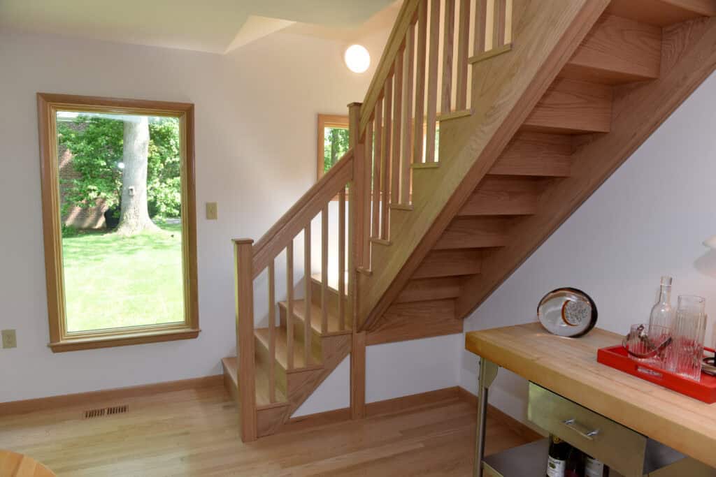 A wooden staircase with vertical railings sits beside a large window overlooking a green yard. Sunlight brightens the room, which has light hardwood floors and a small countertop with glassware on the right.